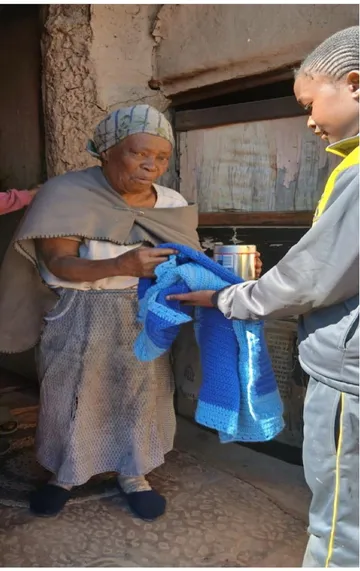 One of our youth handing a hand-knitted blanket and a tin to an elderly woman at her front door.