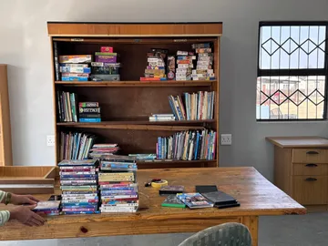 A wooden shelf unit labelled for games and puzzles, with books and donated DVDs stacked on the table in front of it.