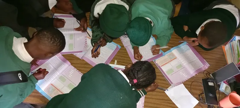 Children in green school tracksuits gathered around a table, heads down over their workbooks.