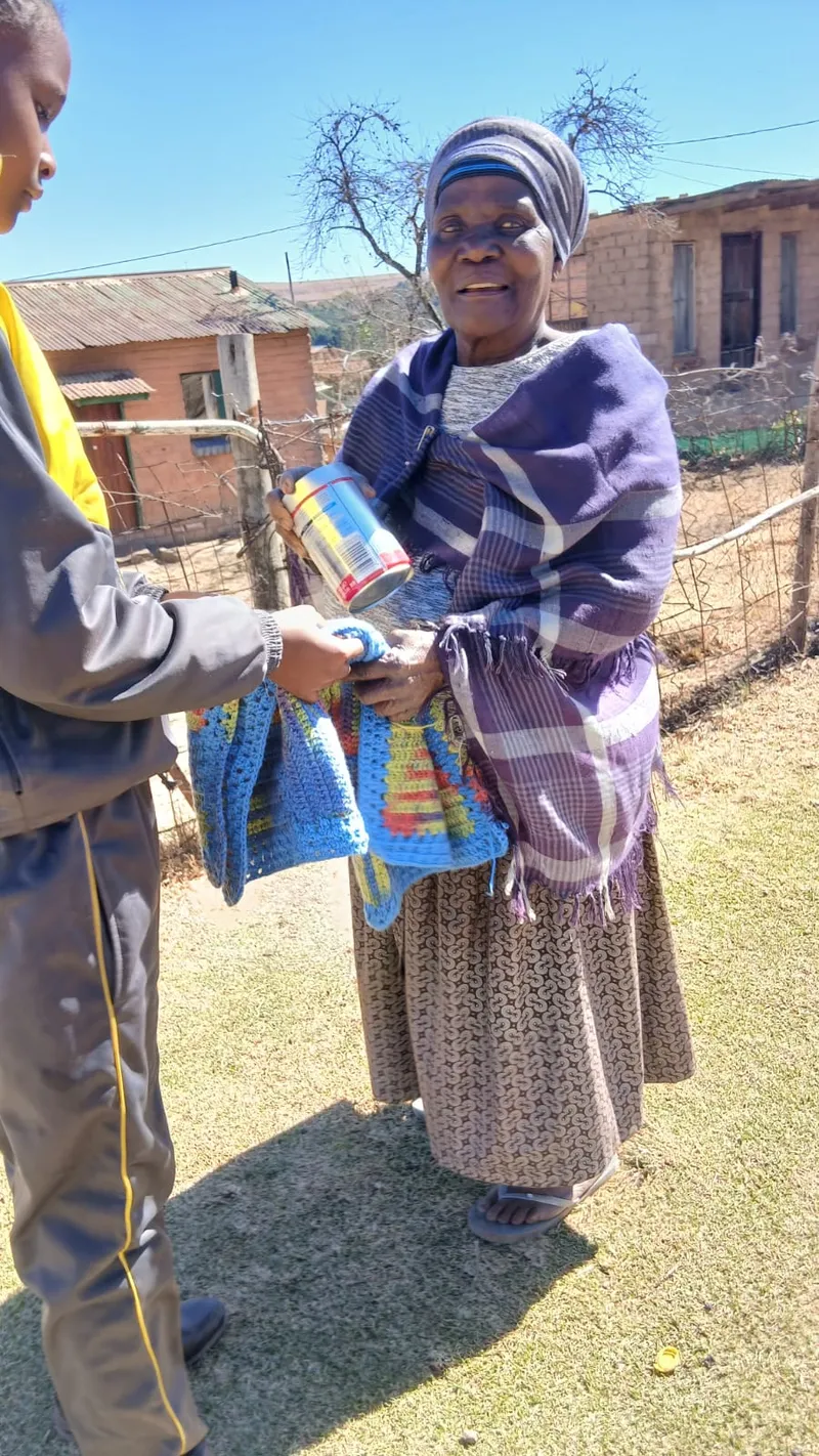 A child handing a knitted blanket and a tin to an elderly woman standing outside her home in the sun.