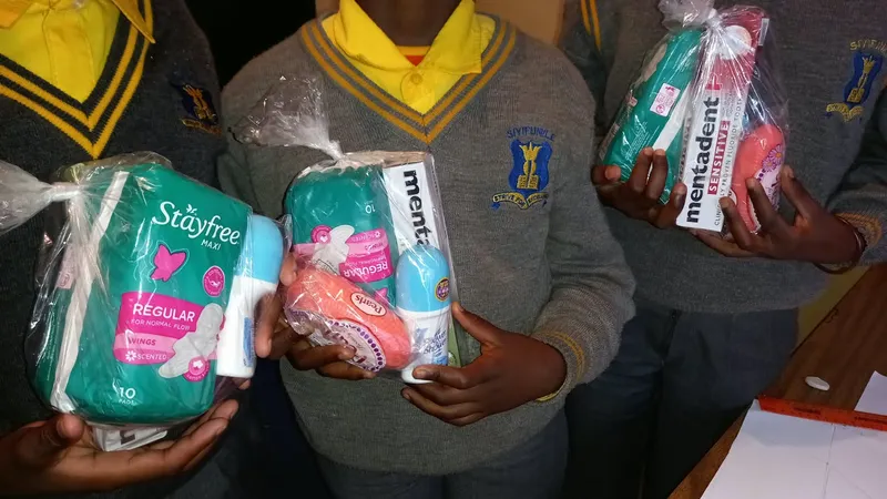 Three children in school jerseys each holding a wrapped pack of pads, soap, roll-on and toothpaste.